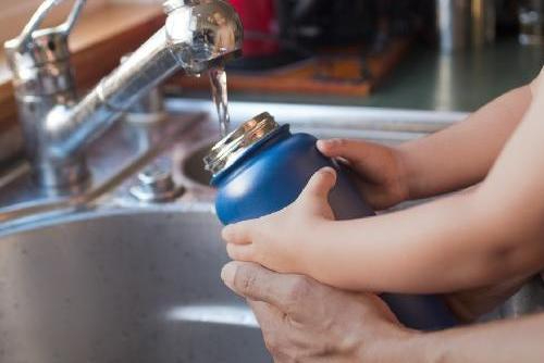 Hands of a child and man filling up a stainless steel water bottle at the kitchen sink. 