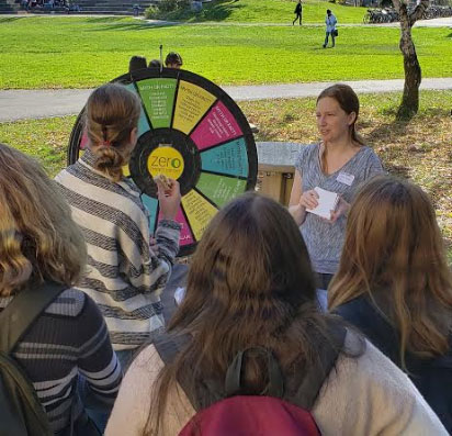 Lianna Hartmour stands next to a wheel that has questions about breast cancer on it. Four students participate in answering the question.