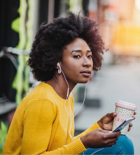 Black woman with a smartphone and coffee wearing headphones.